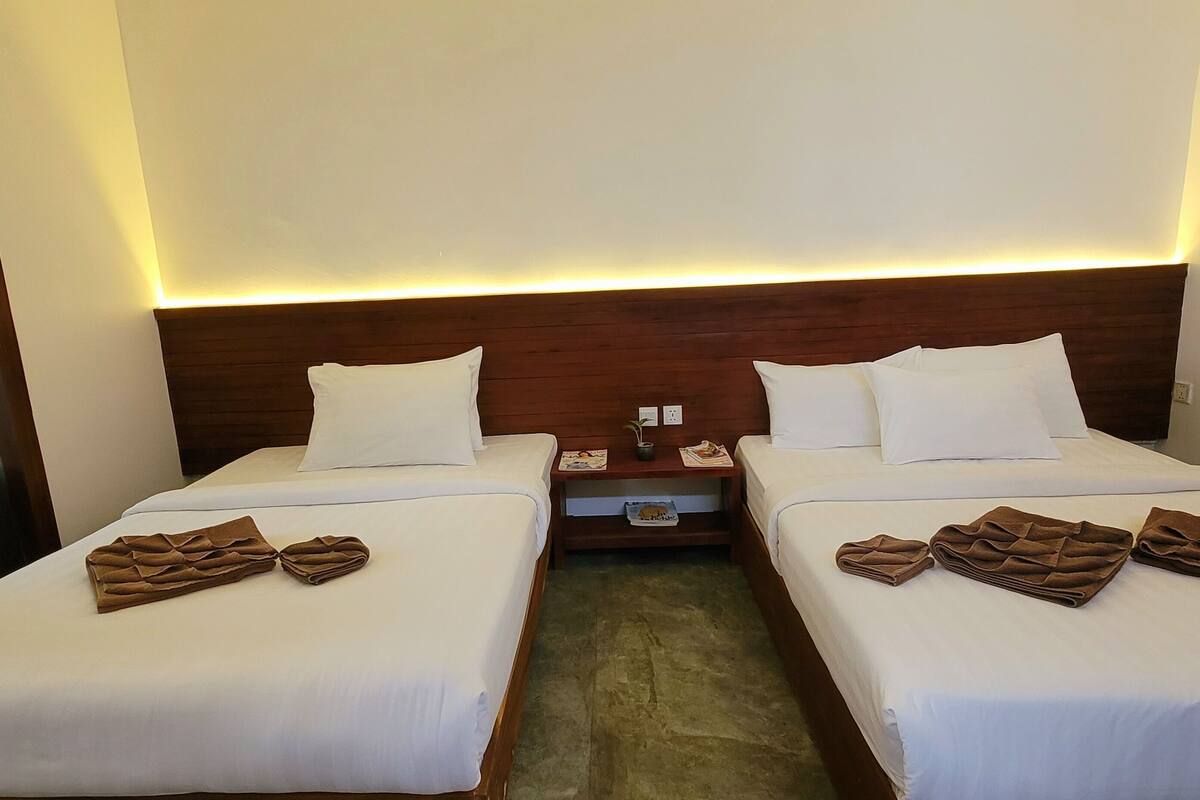 Ein Hotelzimmer mit zwei Betten, jeweils mit weißen Laken und braunen Handtüchern. Es gibt ein hölzernes Kopfteil, einen Nachttisch mit einer Lampe und einen kleinen Tisch mit einer Pflanze.