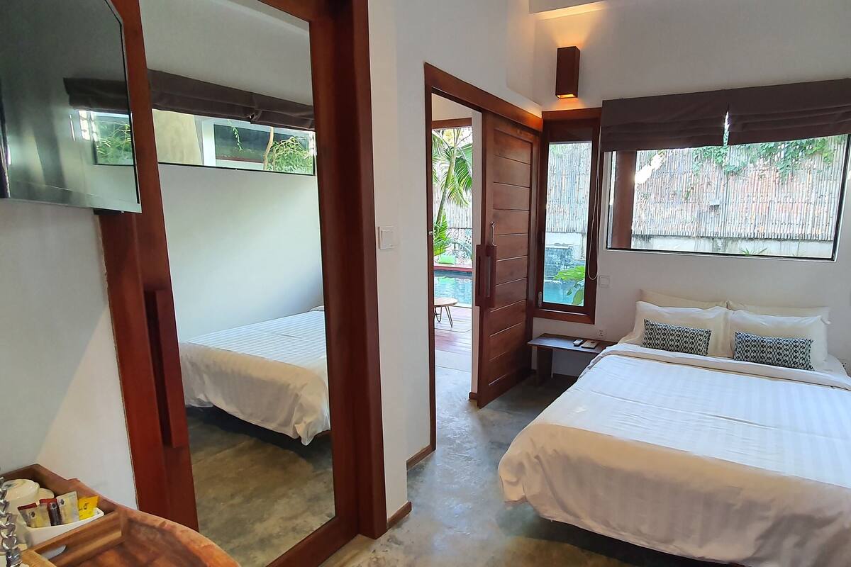 Ein Schlafzimmer mit zwei Betten, einem Fernseher, einem Holzschrank und einem Fenster mit Blick auf einen Pool und Grünflächen.