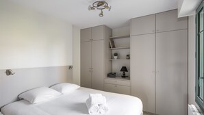 Ein ordentlich bezogenes Bett mit weißen Leinen und aufgerollten Handtüchern, ein Nachttisch mit Lampe sowie ein Kleiderschrank mit offenen Regalen.