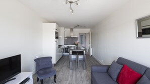 Ein modernes Wohnzimmer mit einem Flachbildfernseher, einem grauen Sofa und einem roten Kissen. Im Hintergrund ist ein Essbereich mit Tisch und Stühlen zu sehen.