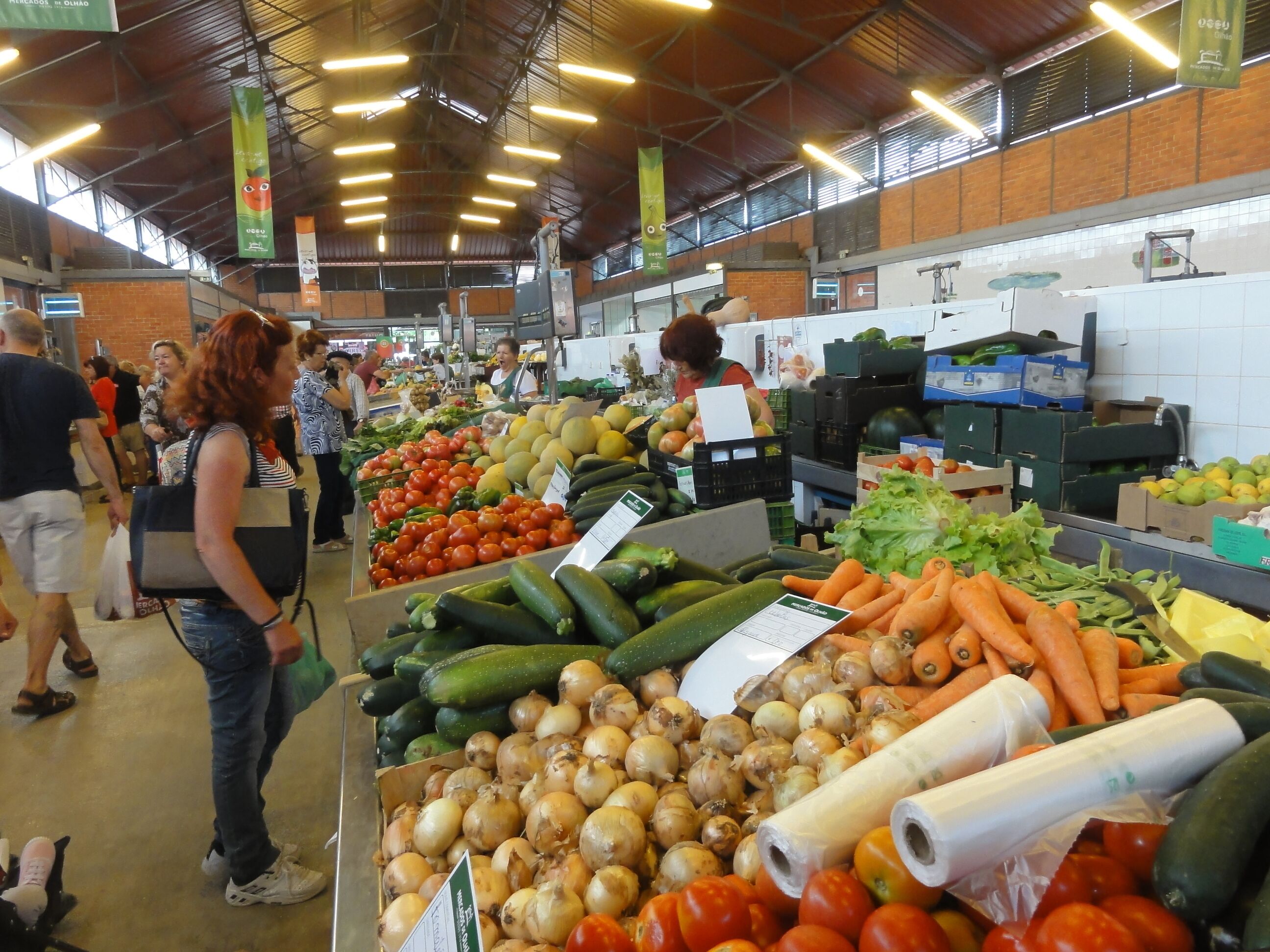A bustling indoor market with various fresh produce on display, including tomatoes, cucumbers, and onions.