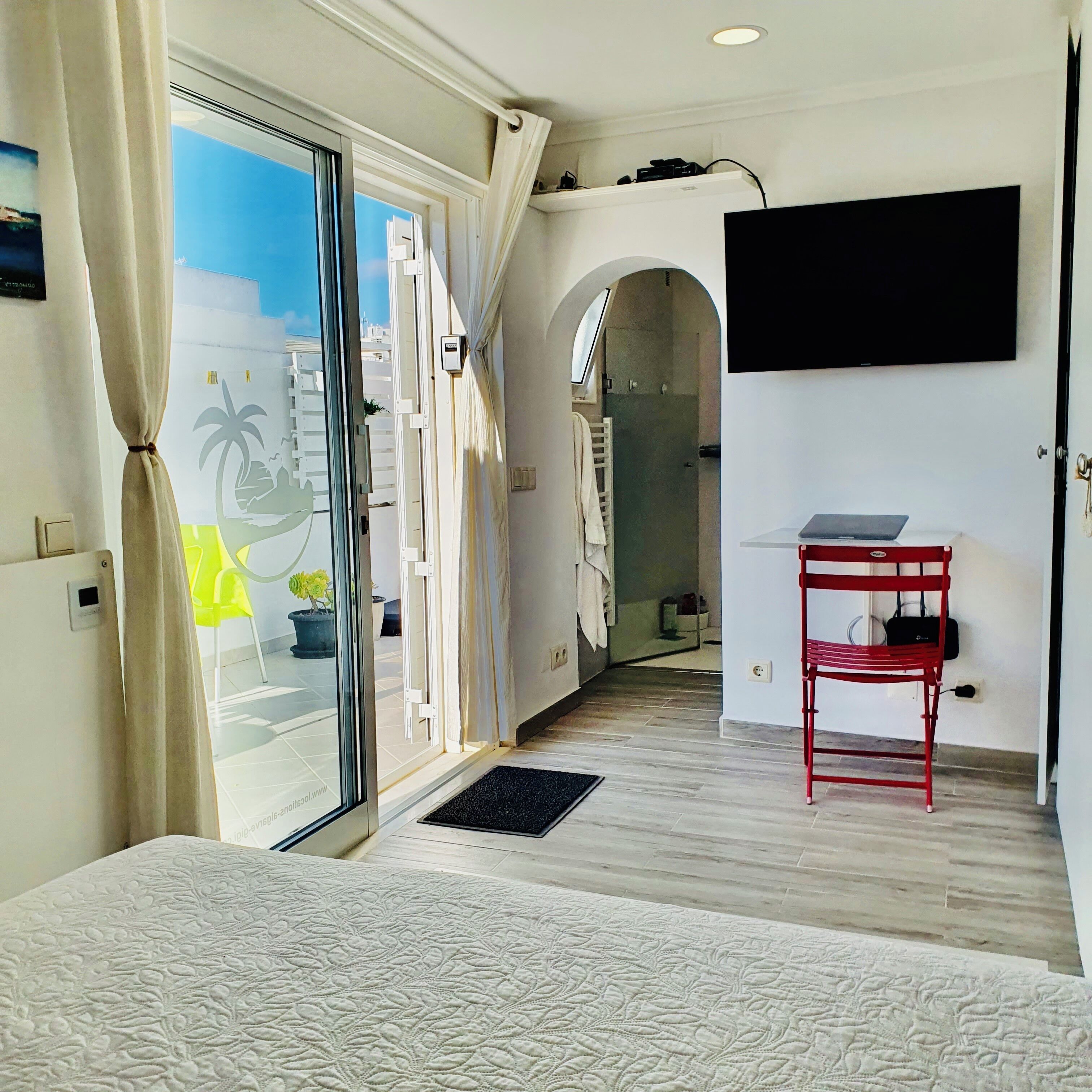 Une chambre d’hôtel avec un lit, un téléviseur fixé au mur, un fauteuil rouge et une vue sur l’extérieur par une porte vitrée.