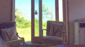 A room with a large sliding glass door leading to a balcony with a view of the ocean. There are two chairs with decorative pillows.