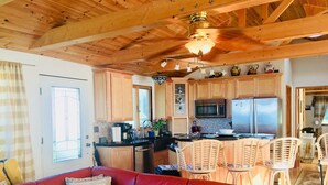 A cozy living room with a red leather sofa, wooden ceiling beams, and a kitchen area with wooden cabinets and a stainless steel refrigerator.