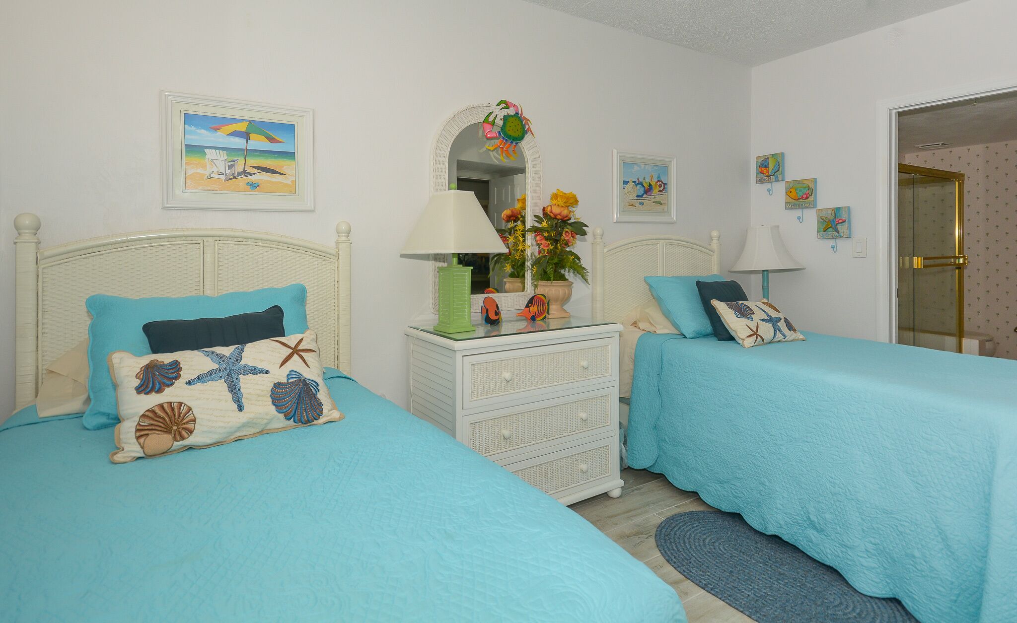 Ein Schlafzimmer mit zwei Betten, jeweils mit blauer Bettdecke und passenden Kissen. Es gibt eine weiße Kommode mit Spiegel, eine Lampe und eine Vase mit Blumen. Das Zimmer ist mit einem Strandbild an der Wand und einem kleinen Fenster mit Vorhängen ausgestattet.