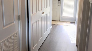 Un pasillo con puertas blancas, una rejilla de ventilación a la izquierda y una puerta con ventanal al final.