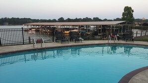 Una piscina con un cartel de prohibición de clavados, rodeada por una valla, y un área techada con sillas y mesas.