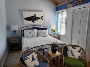 Une chambre à coucher comprenant un lit, deux tables de chevet, une fenêtre avec des stores, une plante en pot et deux fauteuils en osier avec des coussins.