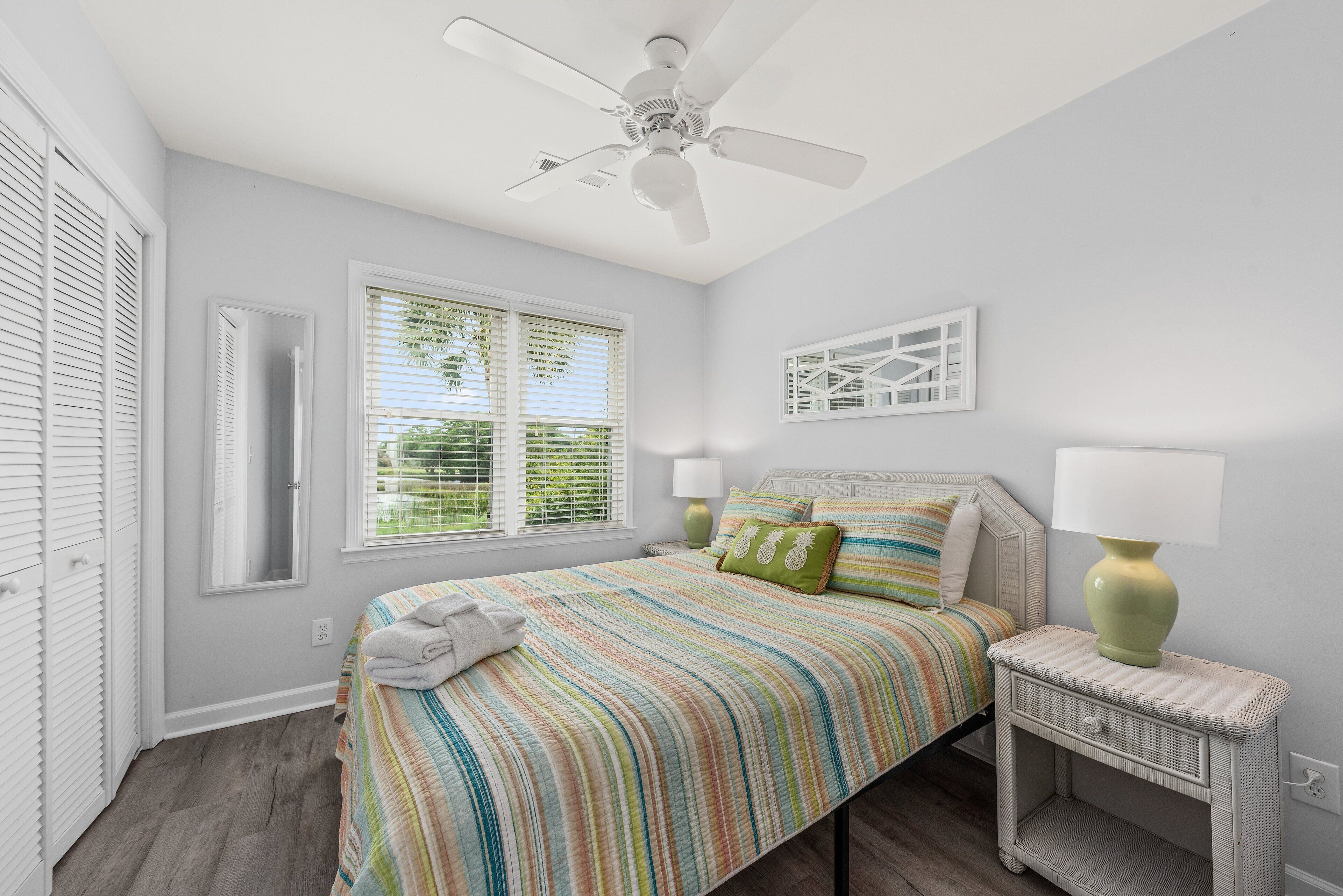 Une chambre à coucher comprenant un lit, une table de chevet, un ventilateur de plafond, une fenêtre avec des stores et un luminaire fixé au mur.