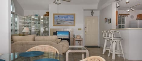 Ein Wohnzimmer mit Glastisch, Korbstühlen, einer Couch, einem Fernseher und einem Deckenventilator.