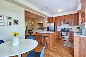 A modern kitchen with wooden cabinets, a stainless steel refrigerator, and a dining area with blue chairs and a white table.