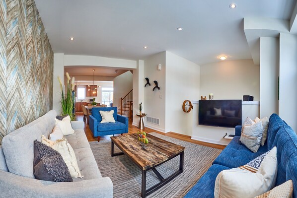 A modern living room with a grey sofa, a blue sofa, and a wooden coffee table. There is a TV on the wall and a staircase leading to another room.
