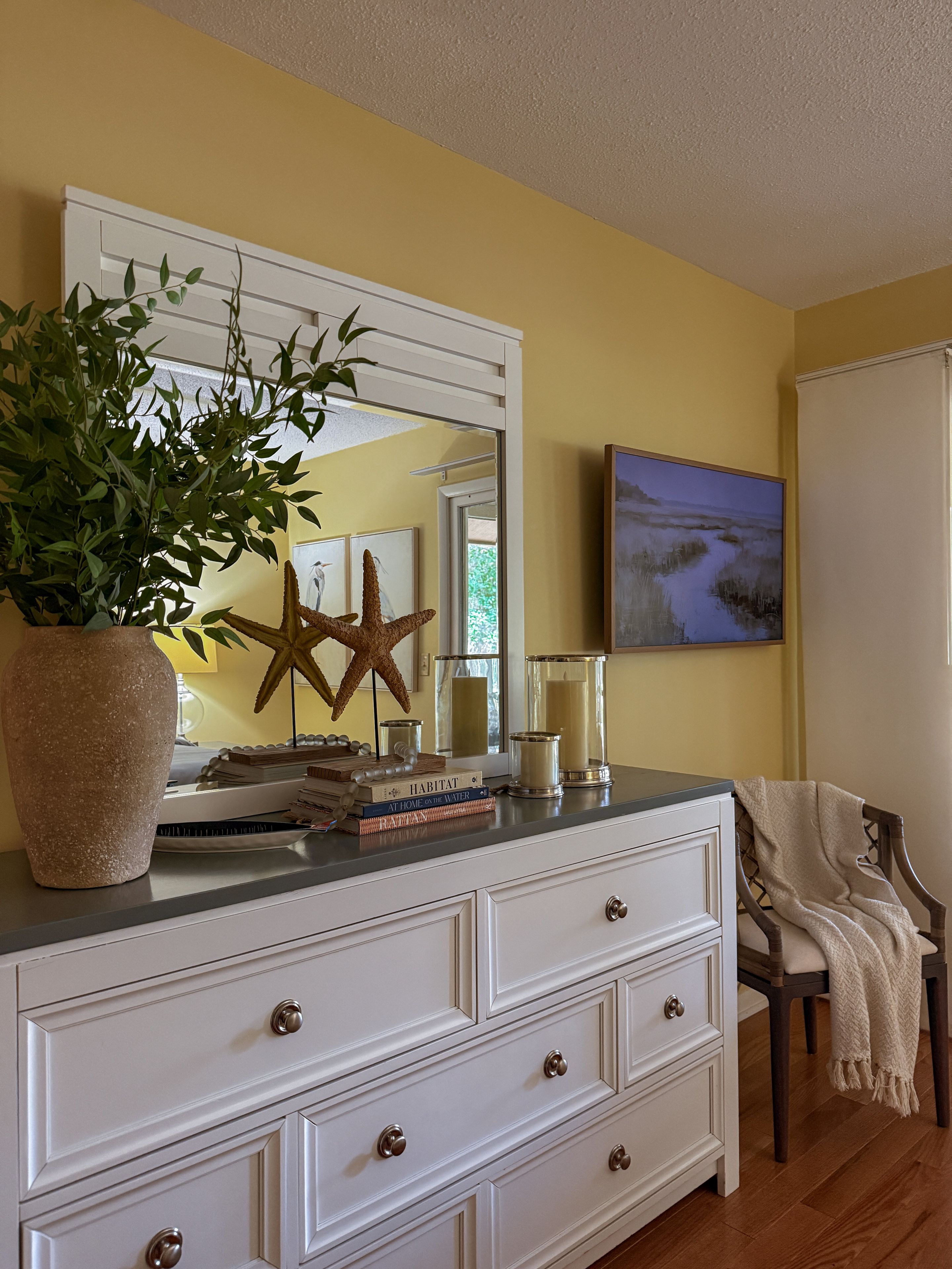 Una habitación con un aparador blanco, una planta en maceta, una decoración de estrella de mar y un cuadro en la pared.