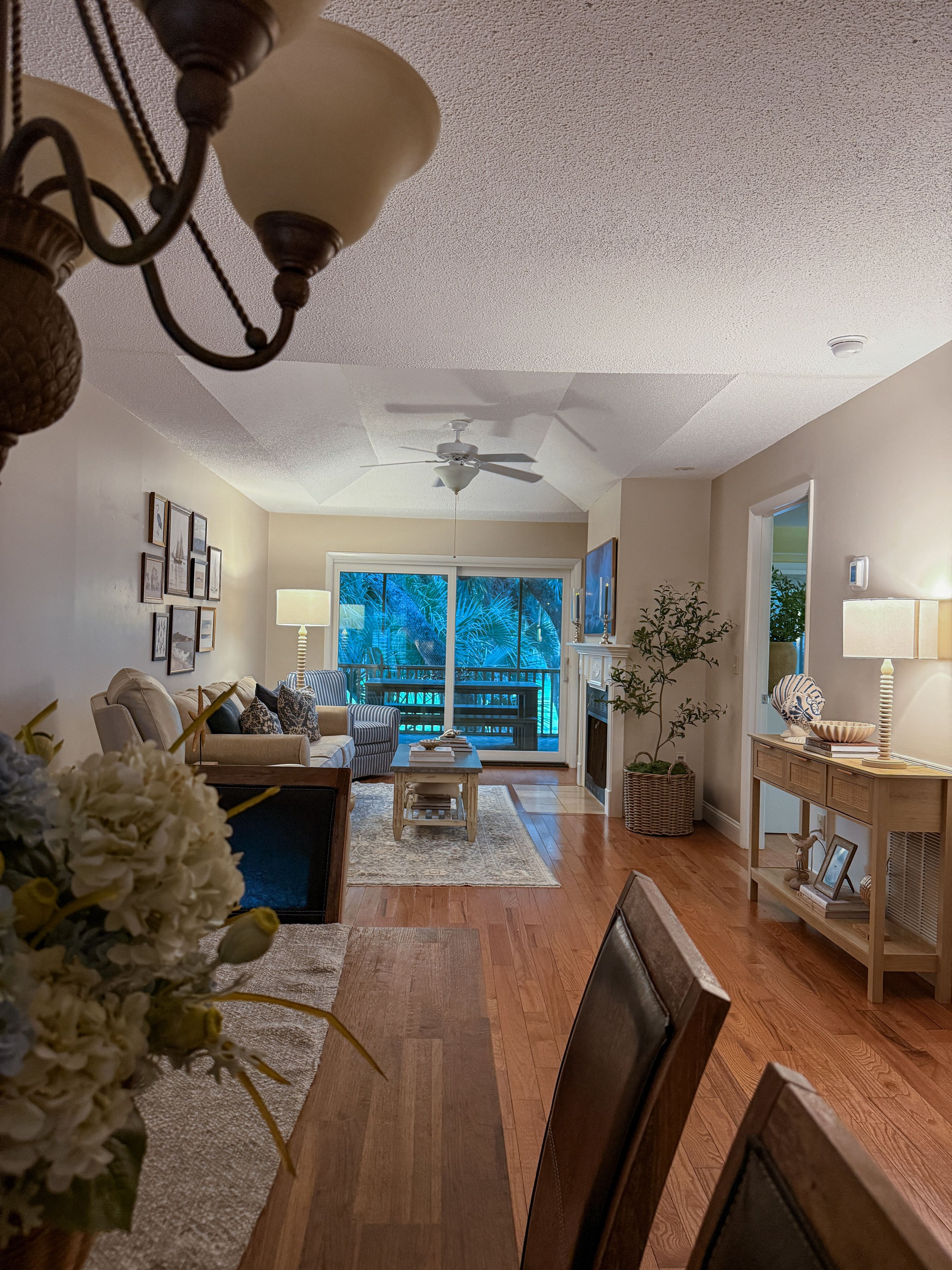 A living room with a ceiling fan, a dining table with chairs, a sofa, and a view of a balcony with plants.