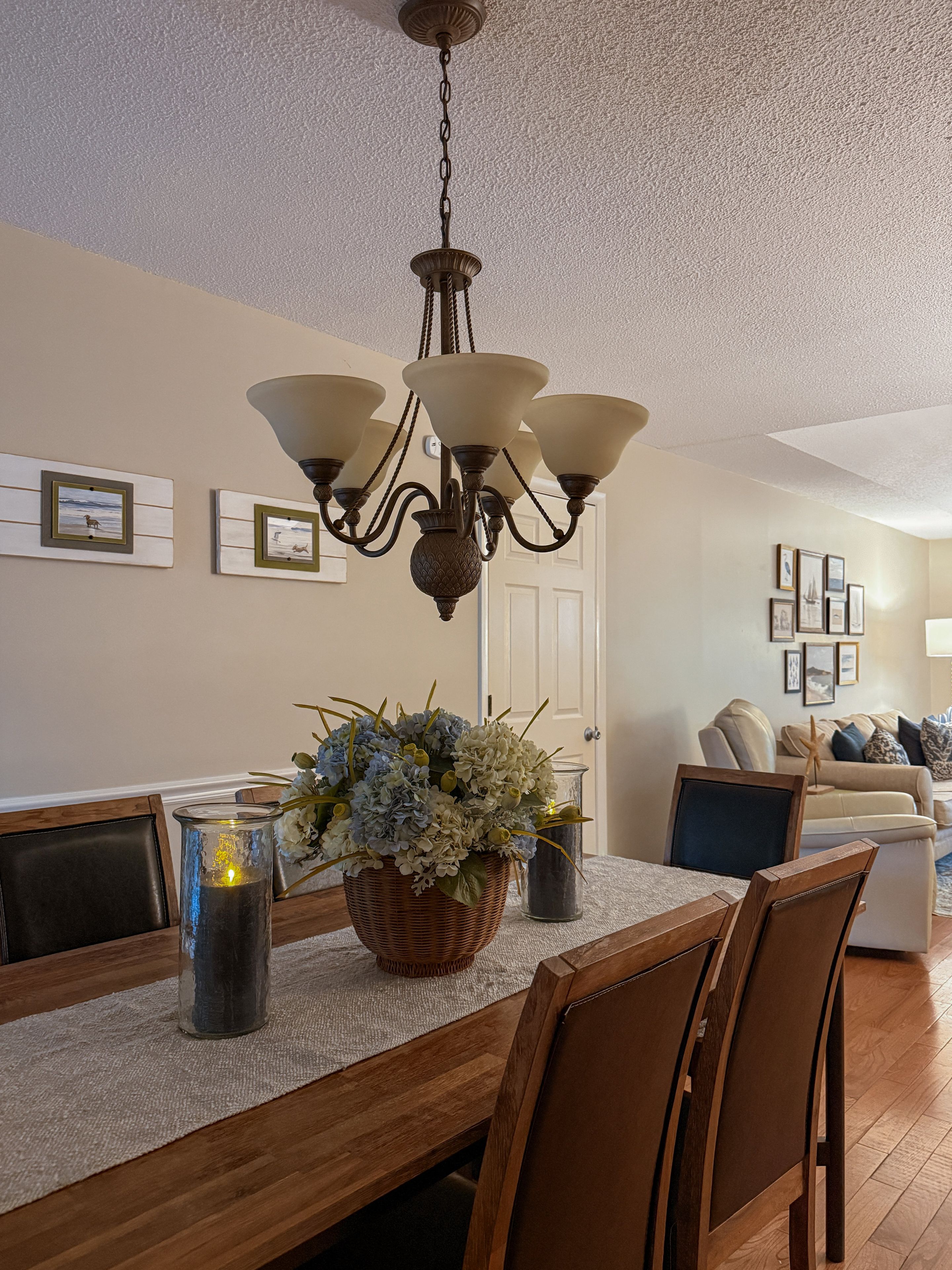 A dining area with a chandelier, a table with chairs, a vase with flowers, and framed pictures on the wall.