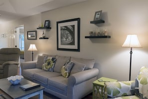 Un salon avec un canapé gris, un fauteuil vert, une petite table sur laquelle se trouvent un bol et un livre, et deux lampadaires.