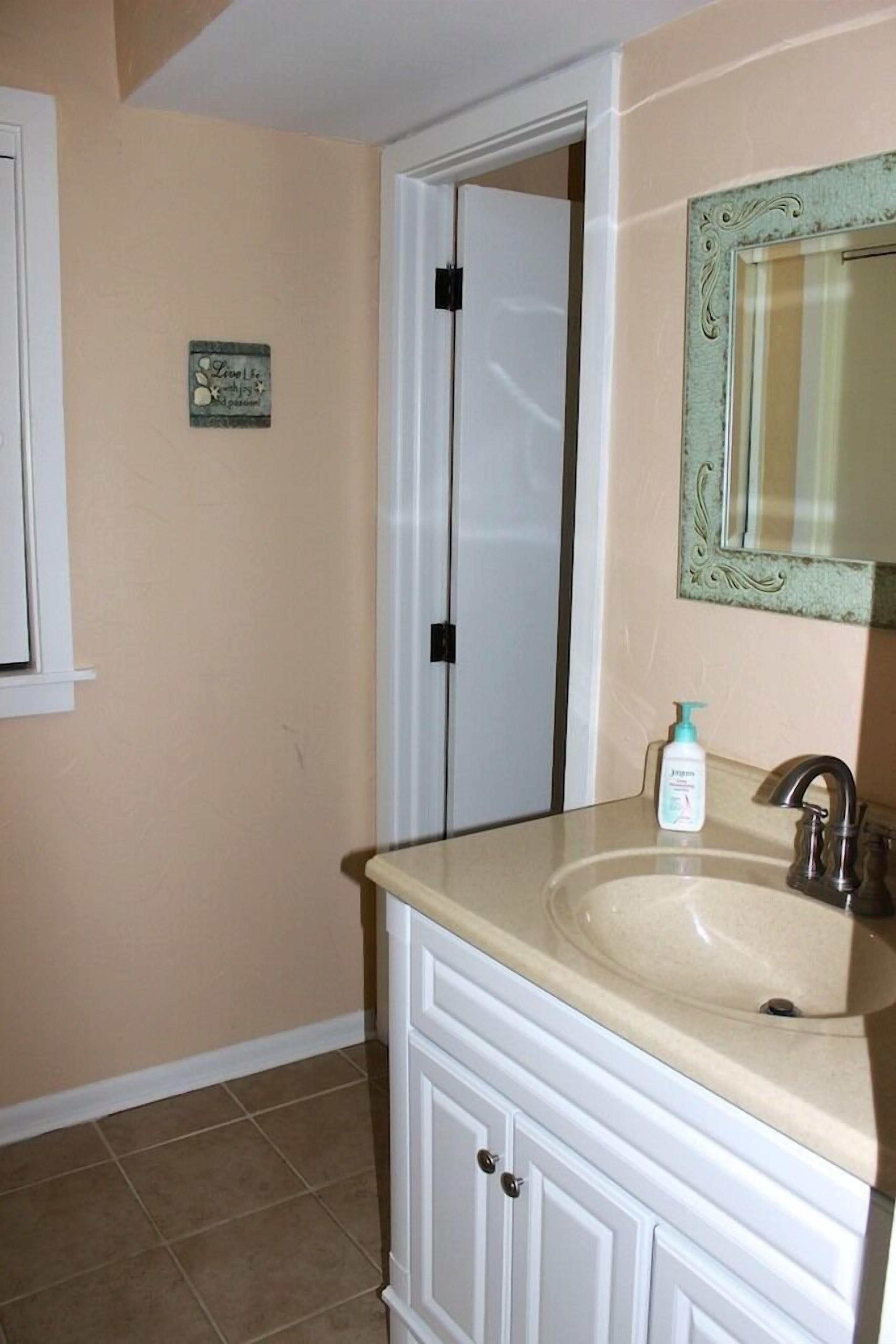 Un baño con pared color crema, una puerta blanca, un lavamanos con espejo y un pequeño letrero decorativo.