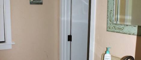 Un baño con pared color crema, una puerta blanca, un lavamanos con espejo y un pequeño letrero decorativo.