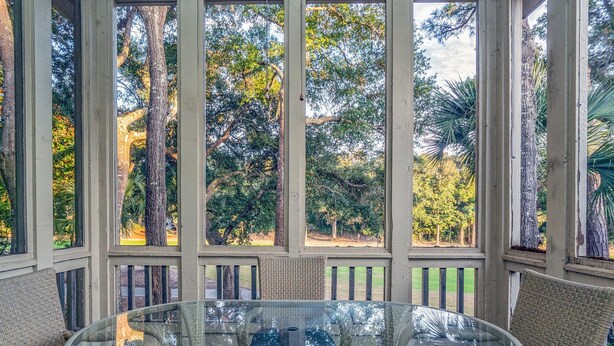 Un espacio de estar al aire libre cubierto, con mobiliario de mimbre y una mesa de vidrio, con vistas a un jardín con árboles y césped.