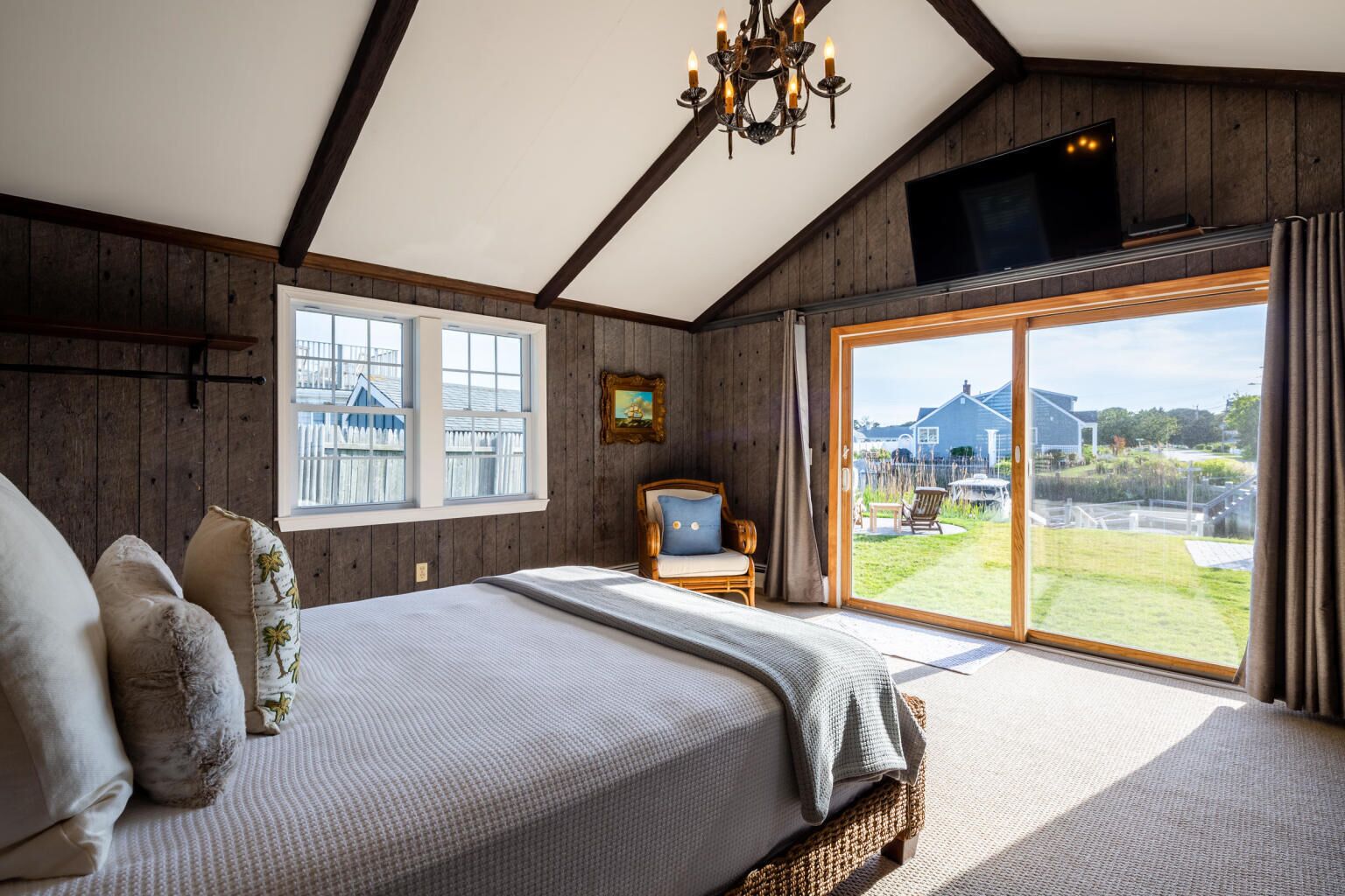 Un dormitorio con una cama grande, televisión, ventana con vista a una casa y vegetación, y un candelabro.