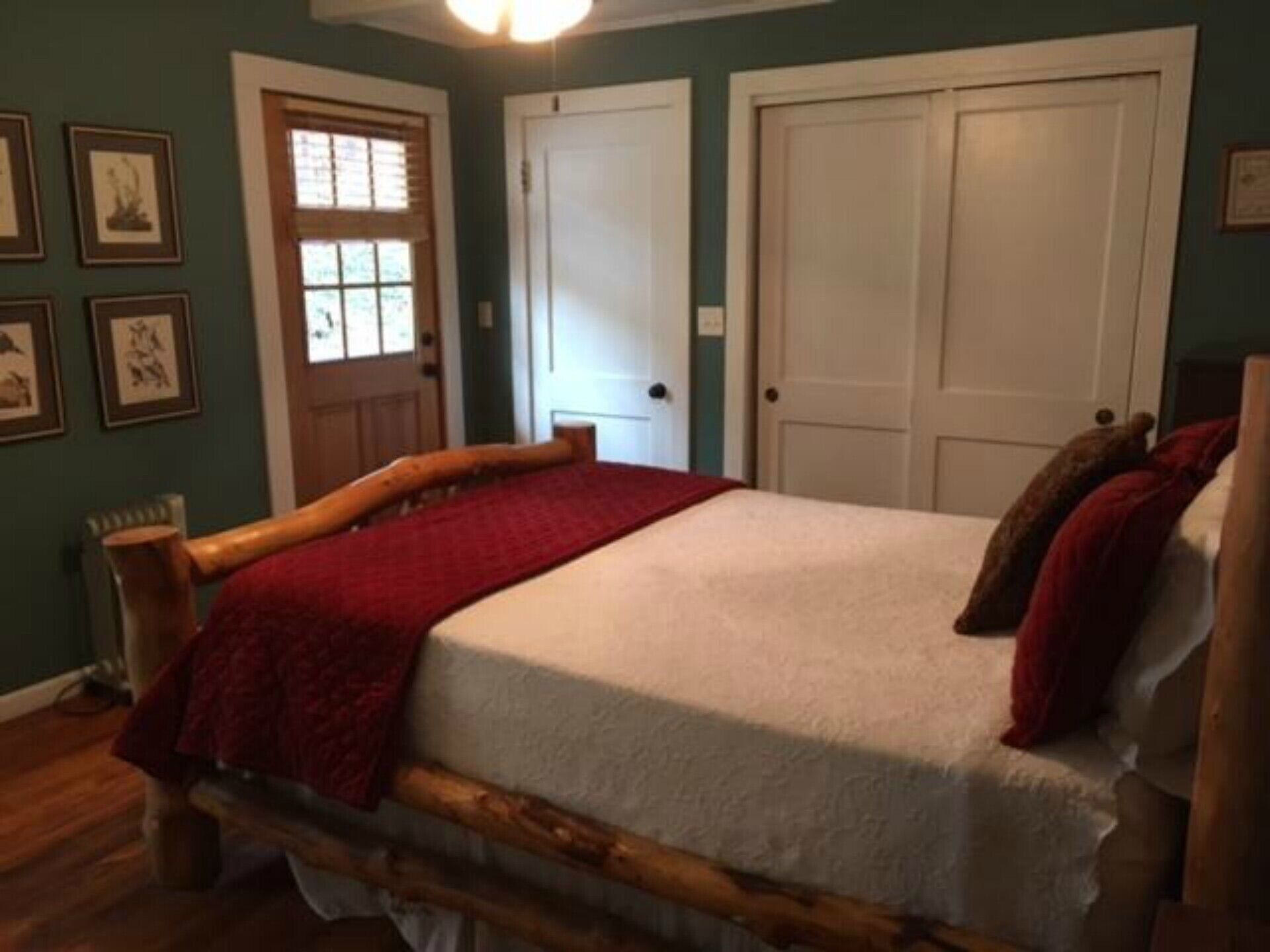 A bedroom with a wooden bed, a red blanket, and a white bedspread. There are two white doors and a window with blinds.