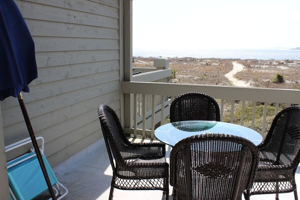 A balcony with wicker chairs and a glass table, overlooking a beach and dunes.