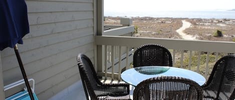 A balcony with wicker chairs and a glass table, overlooking a beach and dunes.