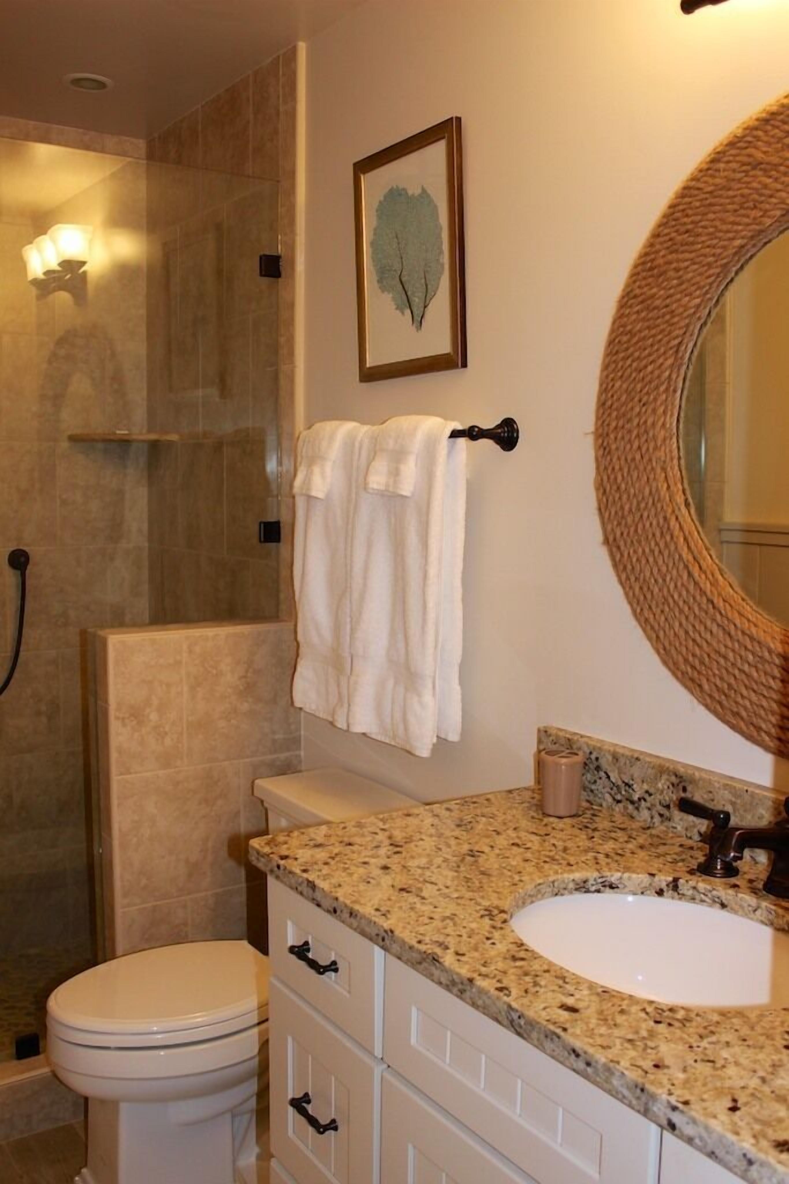Une salle de bain avec des toilettes blanches, un lavabo sur un plan de travail en granit, un grand miroir et un espace douche.