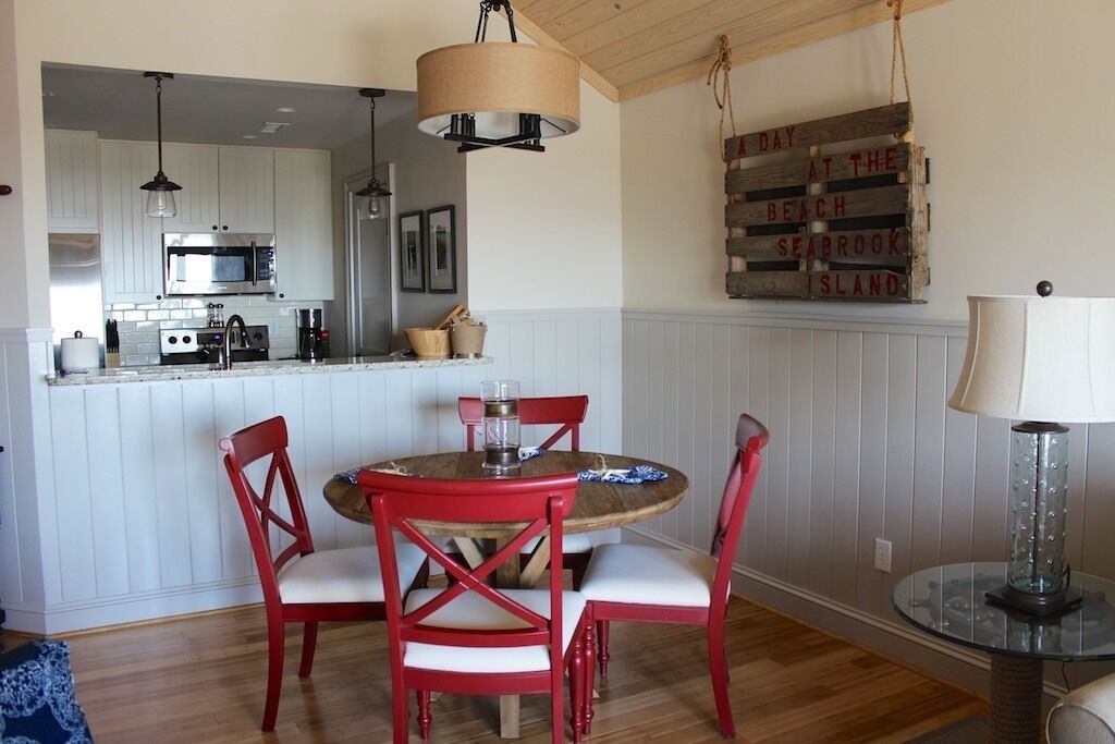 A dining area with a round table, four red chairs, a hanging light fixture, and a wooden sign on the wall.
