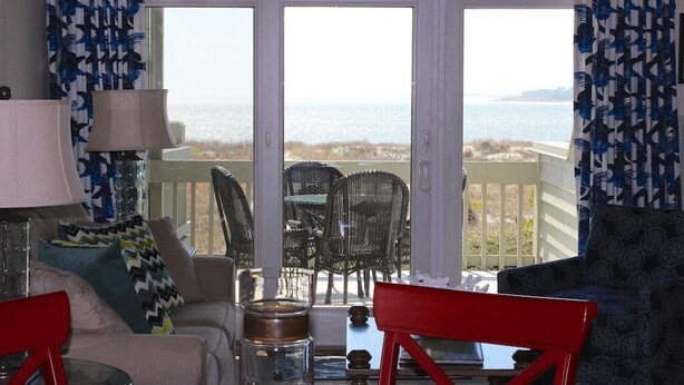 Un salon avec vue sur la mer, un fauteuil rouge, un canapé bleu à motifs et une table avec une lampe et un bocal en verre.