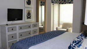 Une chambre à coucher comprenant un lit, une commode avec des tiroirs, un téléviseur, un miroir et une fenêtre avec des stores.