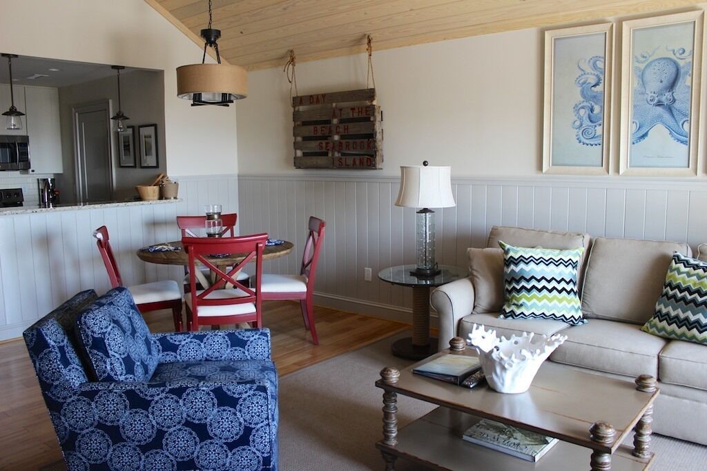 A cozy living room with a sofa, a coffee table, and a red chair. There is a kitchen area in the background with a hanging sign and framed artwork on the wall.