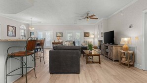 A spacious living room with a sofa, chairs, a coffee table, and a television. There is a dining area with chairs and a table. The room has a ceiling fan and a large window.