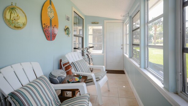 A sunroom with light blue walls, white furniture, and a large window overlooking a lawn. - Charming home looks out onto Hidden Lakes Golf Course. 1152F (New Smyrna Beach)