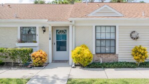 A two-story house with a front porch, a garage door, and a small garden with yellow and green shrubs. - Charming home looks out onto Hidden Lakes Golf Course. 1152F (New Smyrna Beach)