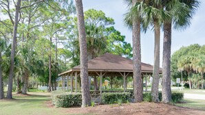 A gazebo with a wooden roof and open sides, surrounded by tall palm trees and other greenery. - Charming home looks out onto Hidden Lakes Golf Course. 1152F (New Smyrna Beach)