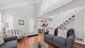 Amplia sala de estar con un sofá gris, un sillón a rayas y una mesa de centro redonda. Una escalera con barandillas blancas conduce a un nivel superior.