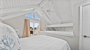 Un dormitorio con techo inclinado, una cama con colcha blanca acolchada, una ventana con cortinas y un armario blanco.