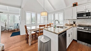 Una cocina moderna con armarios blancos, encimera de granito, electrodomésticos de acero inoxidable y un comedor con sillas de madera y una alfombra azul.