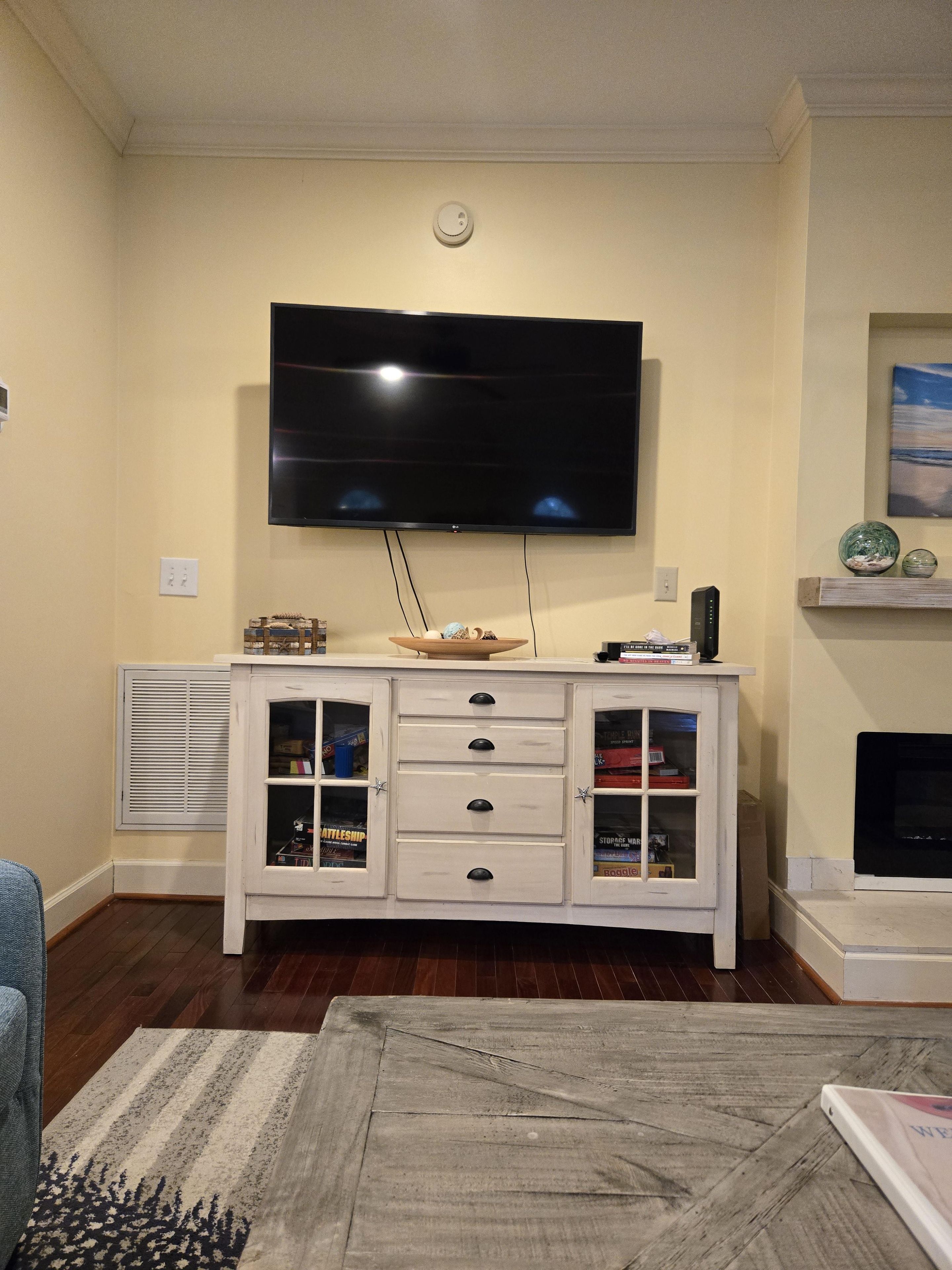 Una sala de estar con un televisor de pantalla plana montado en la pared, un mueble blanco con cajones, una chimenea y una ventana con persianas.
