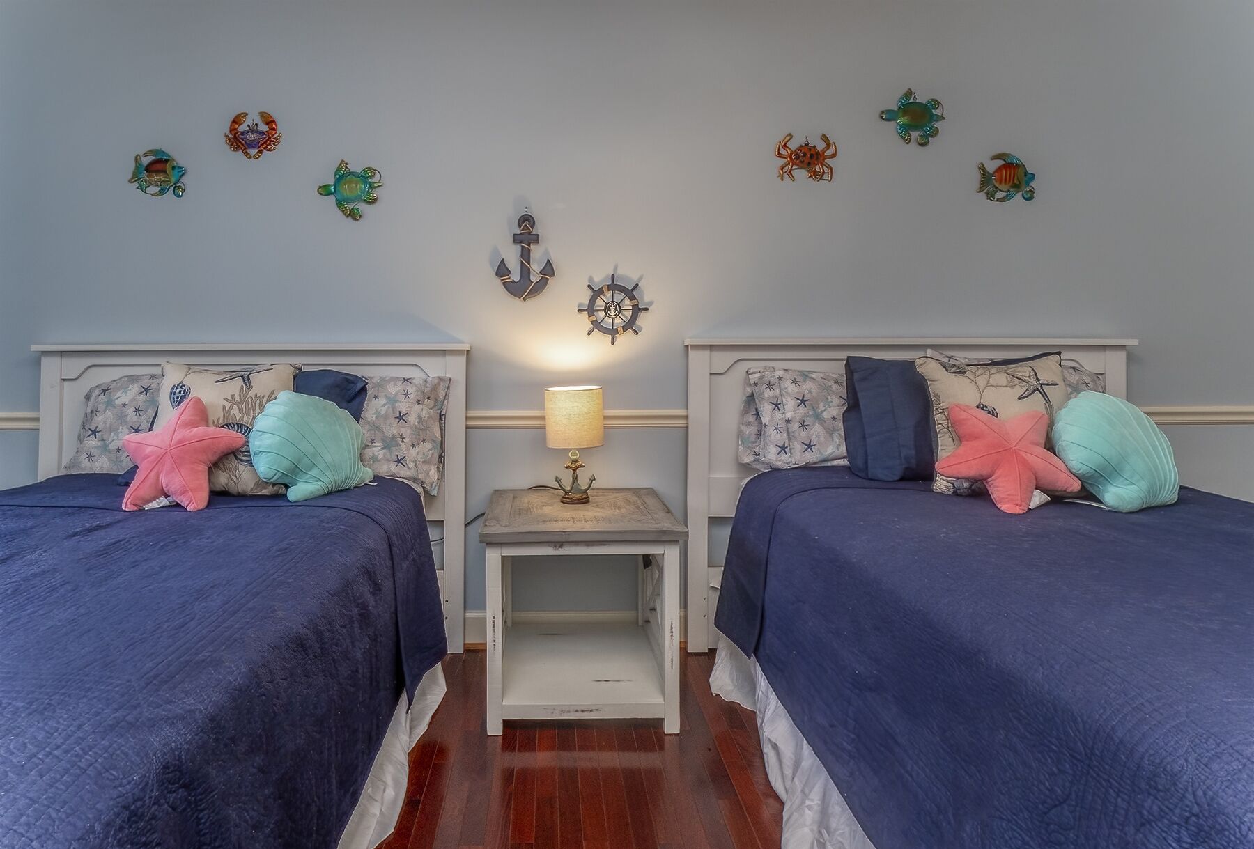 Une chambre avec deux lits, chacun recouvert d’une courtepointe bleue et agrémenté de plusieurs oreillers, une petite table de chevet avec une lampe, et un mur décoré sur le thème de la marine.