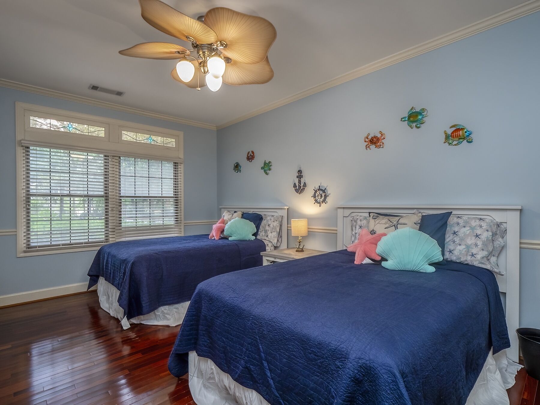 A bedroom with two beds, a ceiling fan, decorative wall art, and a window with blinds.