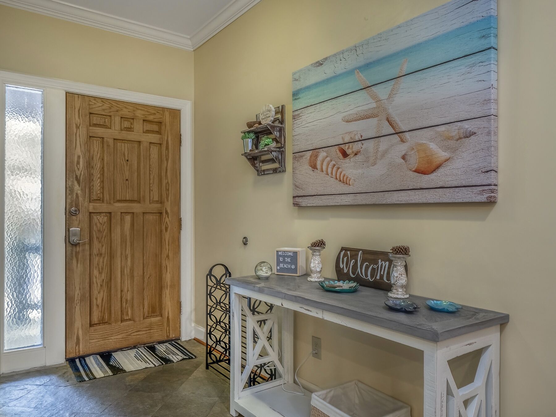 Una porta in legno, uno scaffale a parete con oggetti decorativi, un cartello di benvenuto e un quadro con conchiglie.