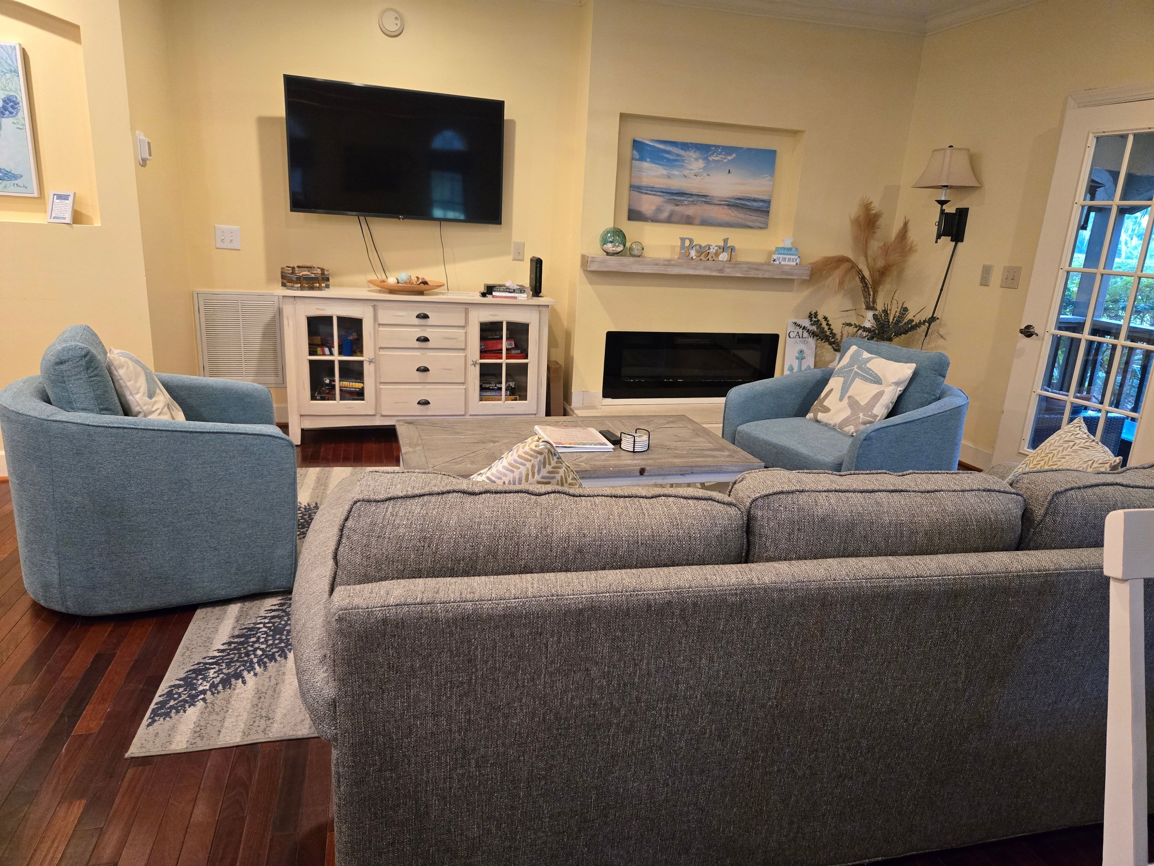 Una sala de estar con un sofá gris, sillones azules, un televisor de pantalla plana, una chimenea y una estantería.