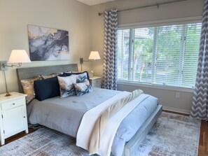 Un dormitorio con cama, mesitas de noche, lámpara, cuadro y ventana con persianas.