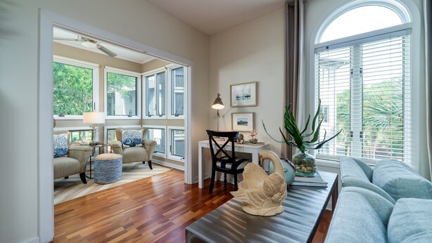 Ein geräumiges Wohnzimmer mit Holzboden, einem großen Fenster mit Jalousien, einem Sofa, einem Esstisch mit Stühlen und einer dekorativen Vase.