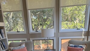 A living room with two patterned armchairs, a round ottoman, a bookshelf, and large windows with a view of trees. - Gorgeous & Sophisticated Villa! Golf Views! Lovely Decor!  Amenities included! Pet Friendly (Seabrook Island)