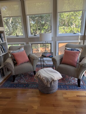 A living room with two patterned armchairs, a round ottoman, a bookshelf, and large windows with a view of trees. - Gorgeous & Sophisticated Villa! Golf Views! Lovely Decor!  Amenities included! Pet Friendly (Seabrook Island)