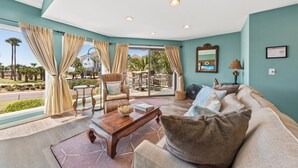 A spacious living room with a view of palm trees and a pool, featuring a sofa, a wooden coffee table, and a wicker chair. - Ocean View 3BR Spinnaker! Steps to Beach, Pools, Dining, Golf! Amenity access included! (Seabrook Island)