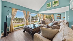 A spacious living room with a large window, a glass coffee table, and a beige sofa. - Ocean View 3BR Spinnaker! Steps to Beach, Pools, Dining, Golf! Amenity access included! (Seabrook Island)